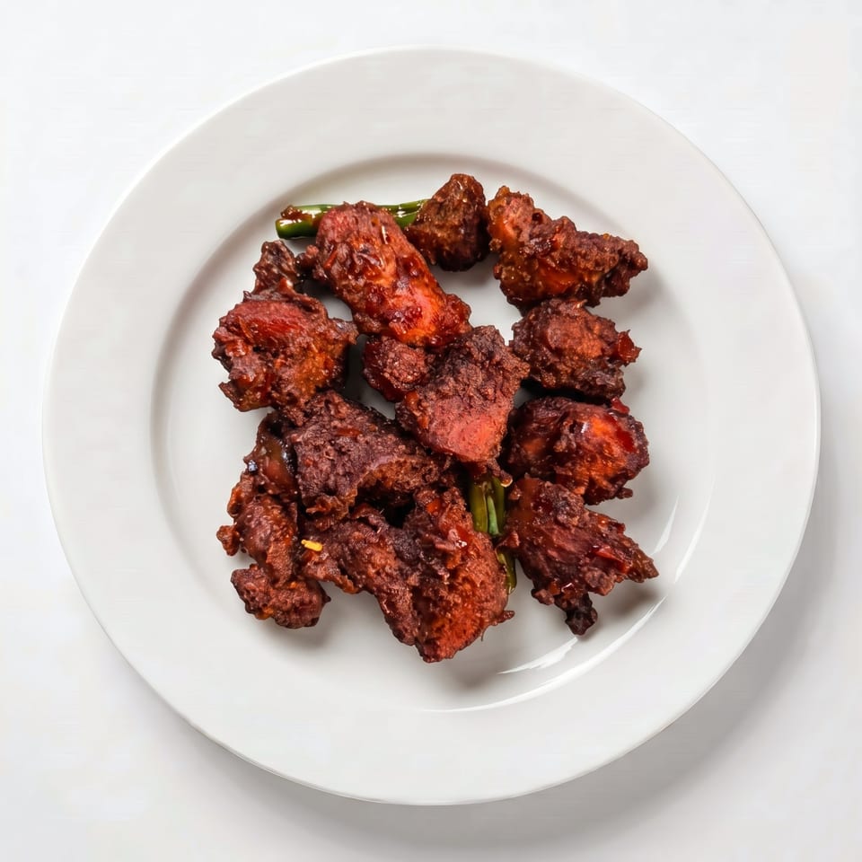 Top-down view of a white plate filled with dark red, crispy fried chicken pieces and green garnishes.